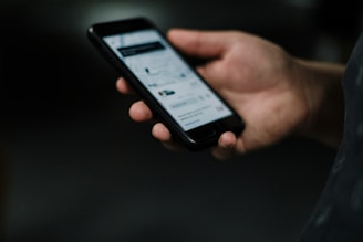 A hand is holding a smartphone with the screen displaying a webpage or app interface, featuring text and images. The background is dark, highlighting the phone.