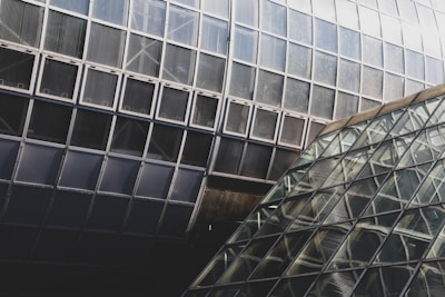 A complex architectural design featuring large geometric glass panes arranged in a grid pattern. The structure shows intersecting angles and reflective surfaces, creating a modern and abstract appearance. The various shades of glass contribute to an intricate visual texture.
