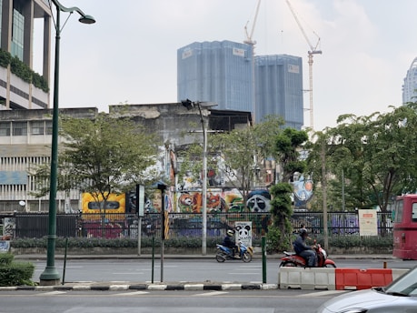 Urban scene featuring a street with motorcycles, trees, a bus, and several buildings. The most prominent building displays vibrant graffiti art, and taller buildings with cranes are visible in the background. There are lamp posts and a fence along the sidewalk.