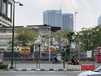 Urban scene featuring a street with motorcycles, trees, a bus, and several buildings. The most prominent building displays vibrant graffiti art, and taller buildings with cranes are visible in the background. There are lamp posts and a fence along the sidewalk.