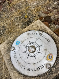 A metal plaque is embedded in the ground with a compass design at the center. The words 'Bukit Pejabat' and 'Jabatan Laut Malaysia' are written around the compass. It features two small emblems and appears to be weathered, surrounded by grass and soil.