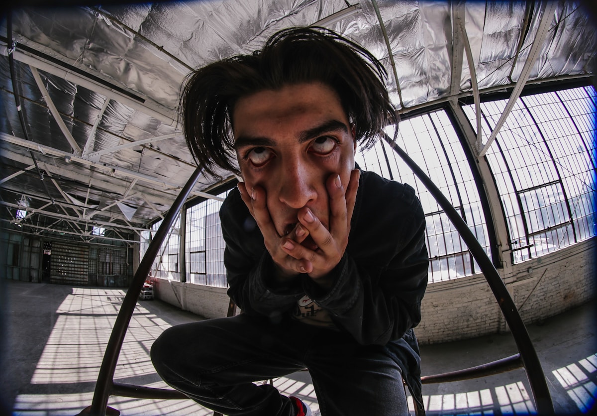 A person in an industrial setting with scaffolding clasping their face, their eyes rolled up into their head.  They wear all black and the view is distorted like a fish bowl.