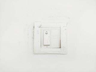 A plain white wall featuring a slightly worn-out light switch panel with a bell symbol on the switch, indicating it might be used for doorbell functions.