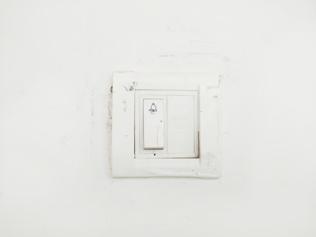 A plain white wall featuring a slightly worn-out light switch panel with a bell symbol on the switch, indicating it might be used for doorbell functions.