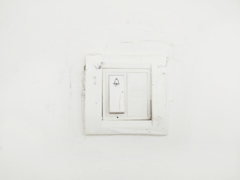 A plain white wall featuring a slightly worn-out light switch panel with a bell symbol on the switch, indicating it might be used for doorbell functions.