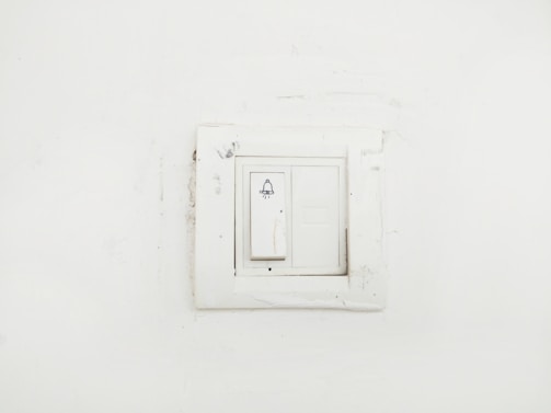 A plain white wall featuring a slightly worn-out light switch panel with a bell symbol on the switch, indicating it might be used for doorbell functions.