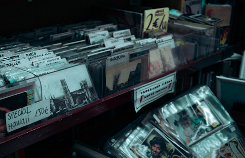 A collection of vintage postcards and photographs of various cities, organized in a retail display. Visible labels indicate locations such as Paris, Brussels, and the United Kingdom. Signs with price tags and promotional offers are scattered among the postcards. The overall arrangement suggests a retro or nostalgic theme.