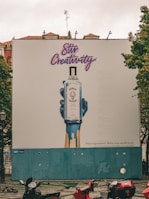 A large outdoor billboard displays an advertisement for Bombay Sapphire gin. The ad features an artistic image of a blue-painted hand holding a bottle of Bombay Sapphire, with blue paint dripping down. Above the image, the text 'Stir Creativity' is written in a stylish, cursive font. Below the image, there is smaller text in Portuguese that advises people to drink responsibly. The background of the billboard is predominantly white with some blue. In the foreground, there are several motorbikes parked on a patterned pavement, and some pigeons can be seen.