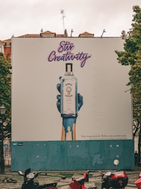 A large outdoor billboard displays an advertisement for Bombay Sapphire gin. The ad features an artistic image of a blue-painted hand holding a bottle of Bombay Sapphire, with blue paint dripping down. Above the image, the text 'Stir Creativity' is written in a stylish, cursive font. Below the image, there is smaller text in Portuguese that advises people to drink responsibly. The background of the billboard is predominantly white with some blue. In the foreground, there are several motorbikes parked on a patterned pavement, and some pigeons can be seen.