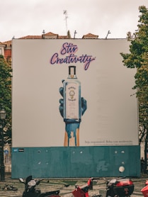 A large outdoor billboard displays an advertisement for Bombay Sapphire gin. The ad features an artistic image of a blue-painted hand holding a bottle of Bombay Sapphire, with blue paint dripping down. Above the image, the text 'Stir Creativity' is written in a stylish, cursive font. Below the image, there is smaller text in Portuguese that advises people to drink responsibly. The background of the billboard is predominantly white with some blue. In the foreground, there are several motorbikes parked on a patterned pavement, and some pigeons can be seen.