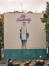 A large outdoor billboard displays an advertisement for Bombay Sapphire gin. The ad features an artistic image of a blue-painted hand holding a bottle of Bombay Sapphire, with blue paint dripping down. Above the image, the text 'Stir Creativity' is written in a stylish, cursive font. Below the image, there is smaller text in Portuguese that advises people to drink responsibly. The background of the billboard is predominantly white with some blue. In the foreground, there are several motorbikes parked on a patterned pavement, and some pigeons can be seen.