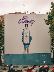 A large outdoor billboard displays an advertisement for Bombay Sapphire gin. The ad features an artistic image of a blue-painted hand holding a bottle of Bombay Sapphire, with blue paint dripping down. Above the image, the text 'Stir Creativity' is written in a stylish, cursive font. Below the image, there is smaller text in Portuguese that advises people to drink responsibly. The background of the billboard is predominantly white with some blue. In the foreground, there are several motorbikes parked on a patterned pavement, and some pigeons can be seen.