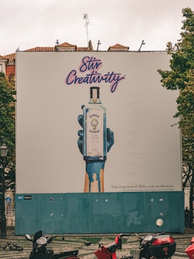 A large outdoor billboard displays an advertisement for Bombay Sapphire gin. The ad features an artistic image of a blue-painted hand holding a bottle of Bombay Sapphire, with blue paint dripping down. Above the image, the text 'Stir Creativity' is written in a stylish, cursive font. Below the image, there is smaller text in Portuguese that advises people to drink responsibly. The background of the billboard is predominantly white with some blue. In the foreground, there are several motorbikes parked on a patterned pavement, and some pigeons can be seen.