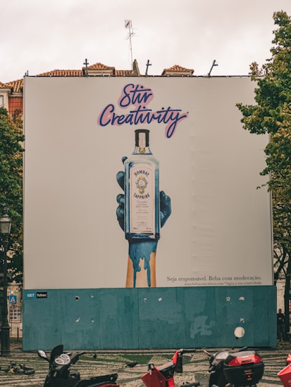 A large outdoor billboard displays an advertisement for Bombay Sapphire gin. The ad features an artistic image of a blue-painted hand holding a bottle of Bombay Sapphire, with blue paint dripping down. Above the image, the text 'Stir Creativity' is written in a stylish, cursive font. Below the image, there is smaller text in Portuguese that advises people to drink responsibly. The background of the billboard is predominantly white with some blue. In the foreground, there are several motorbikes parked on a patterned pavement, and some pigeons can be seen.