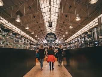 A spacious, modern market hall with a high ceiling and industrial lighting. Three people are walking through the aisle, surrounded by various market stalls and products. The space is bustling with activity, featuring a prominent sign reading 'Time Out Market Lisbon'.