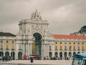 A historic archway, adorned with elaborate sculptures and engravings, stands prominently in an open plaza. The surrounding buildings feature bright yellow facades with white detailing and red-tiled roofs. Numerous people are gathered around, and a red tour bus is visible, adding a lively element to the scene. Cloudy skies create a muted backdrop.