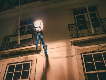 An outdoor street lamp illuminates a residence at night. The light casts warm tones on the walls and balconies, which have ornate iron railings. Blue garlands decorate the area, adding a festive touch. A sign on one of the balconies reads 'STA. MARIA MAIOR'.
