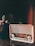 A vintage radio with a retro design is situated next to a potted cactus on a wooden surface. The radio features a metallic finish with a visible tuning scale, and the cactus has small spines and tufts of pink flowers. A warm, slightly reddish light adds an atmospheric glow to the scene.