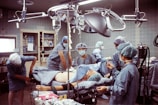 A group of medical professionals are gathered around a patient on an operating table in a hospital room. They are wearing surgical scrubs, masks, and caps, working under bright surgical lights. The room is equipped with various medical instruments including IVs and a monitoring device. The scene conveys a sense of focus and teamwork.