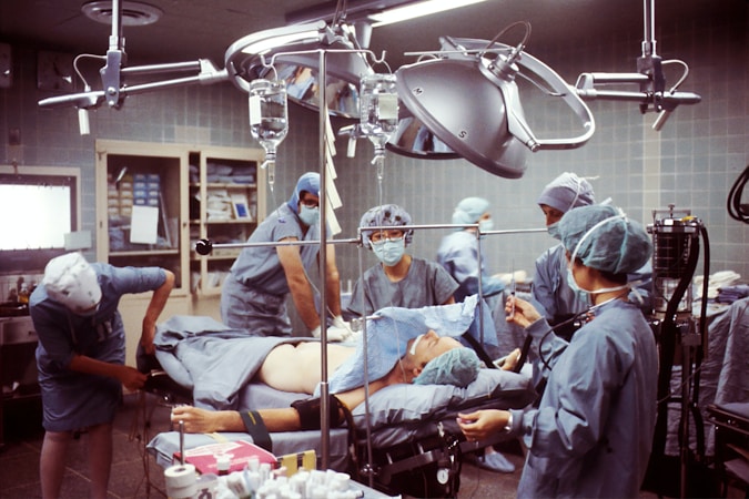 A group of medical professionals are gathered around a patient on an operating table in a hospital room. They are wearing surgical scrubs, masks, and caps, working under bright surgical lights. The room is equipped with various medical instruments including IVs and a monitoring device. The scene conveys a sense of focus and teamwork.