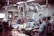 A group of medical professionals are gathered around a patient on an operating table in a hospital room. They are wearing surgical scrubs, masks, and caps, working under bright surgical lights. The room is equipped with various medical instruments including IVs and a monitoring device. The scene conveys a sense of focus and teamwork.