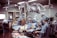 A group of medical professionals are gathered around a patient on an operating table in a hospital room. They are wearing surgical scrubs, masks, and caps, working under bright surgical lights. The room is equipped with various medical instruments including IVs and a monitoring device. The scene conveys a sense of focus and teamwork.