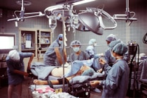 A group of medical professionals are gathered around a patient on an operating table in a hospital room. They are wearing surgical scrubs, masks, and caps, working under bright surgical lights. The room is equipped with various medical instruments including IVs and a monitoring device. The scene conveys a sense of focus and teamwork.