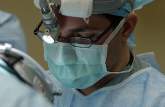 A surgeon wearing a surgical mask and headlight is focused on conducting a medical procedure. The individual is dressed in surgical attire and is working in a sterile environment.