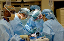 Four surgeons in blue scrubs and masks performing surgery in a bright operating room
