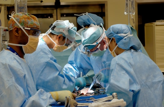 Several surgeons, dressed in blue surgical gowns and protective masks, are performing a medical procedure in an operating room. Surgical instruments, such as scalpels and forceps, are visible in the foreground. The environment is clinical and filled with medical equipment.