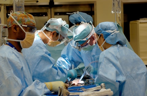 Several surgeons, dressed in blue surgical gowns and protective masks, are performing a medical procedure in an operating room. Surgical instruments, such as scalpels and forceps, are visible in the foreground. The environment is clinical and filled with medical equipment.