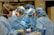 Several surgeons, dressed in blue surgical gowns and protective masks, are performing a medical procedure in an operating room. Surgical instruments, such as scalpels and forceps, are visible in the foreground. The environment is clinical and filled with medical equipment.