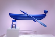 A minimalist art installation featuring a bright blue boat with an oar, balanced precariously on a small blue chair. The setup is displayed on a white platform against a pastel pink backdrop. A balloon-like sphere is attached to the boat.