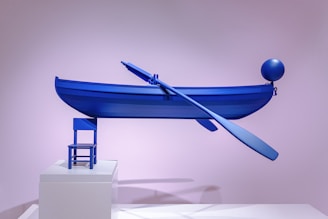 A minimalist art installation featuring a bright blue boat with an oar, balanced precariously on a small blue chair. The setup is displayed on a white platform against a pastel pink backdrop. A balloon-like sphere is attached to the boat.