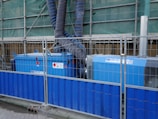 There are industrial machines with blue casings, marked with warning signs. They are surrounded by a metal fence with a blue base. The machines have large black and blue corrugated pipes connected to them, which extend upwards towards green construction netting. This setup suggests a construction or work site environment.