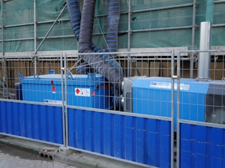 There are industrial machines with blue casings, marked with warning signs. They are surrounded by a metal fence with a blue base. The machines have large black and blue corrugated pipes connected to them, which extend upwards towards green construction netting. This setup suggests a construction or work site environment.