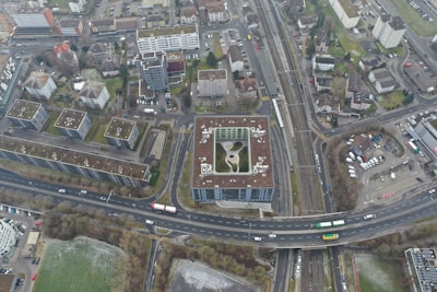 An aerial view of an urban area featuring numerous residential buildings, roads, parking spaces, and a railway line. The buildings exhibit modern architectural designs, predominantly with flat rooftops. A notable structure in the center appears to have a rooftop garden or recreational area. Roads are visible with a few vehicles, including buses and cars, moving through the area. Adjacent to the buildings, there are areas of green space, suggesting the presence of parks or lawns.