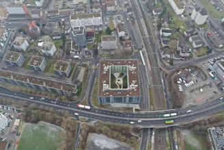 An aerial view of an urban area featuring numerous residential buildings, roads, parking spaces, and a railway line. The buildings exhibit modern architectural designs, predominantly with flat rooftops. A notable structure in the center appears to have a rooftop garden or recreational area. Roads are visible with a few vehicles, including buses and cars, moving through the area. Adjacent to the buildings, there are areas of green space, suggesting the presence of parks or lawns.