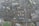 An aerial view of an urban area featuring numerous residential buildings, roads, parking spaces, and a railway line. The buildings exhibit modern architectural designs, predominantly with flat rooftops. A notable structure in the center appears to have a rooftop garden or recreational area. Roads are visible with a few vehicles, including buses and cars, moving through the area. Adjacent to the buildings, there are areas of green space, suggesting the presence of parks or lawns.