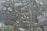 An aerial view of an urban area featuring numerous residential buildings, roads, parking spaces, and a railway line. The buildings exhibit modern architectural designs, predominantly with flat rooftops. A notable structure in the center appears to have a rooftop garden or recreational area. Roads are visible with a few vehicles, including buses and cars, moving through the area. Adjacent to the buildings, there are areas of green space, suggesting the presence of parks or lawns.