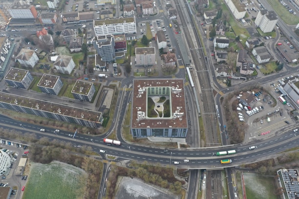 An aerial view of an urban area featuring numerous residential buildings, roads, parking spaces, and a railway line. The buildings exhibit modern architectural designs, predominantly with flat rooftops. A notable structure in the center appears to have a rooftop garden or recreational area. Roads are visible with a few vehicles, including buses and cars, moving through the area. Adjacent to the buildings, there are areas of green space, suggesting the presence of parks or lawns.