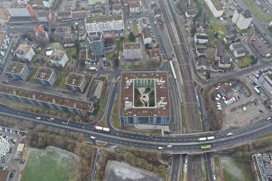 An aerial view of an urban area featuring numerous residential buildings, roads, parking spaces, and a railway line. The buildings exhibit modern architectural designs, predominantly with flat rooftops. A notable structure in the center appears to have a rooftop garden or recreational area. Roads are visible with a few vehicles, including buses and cars, moving through the area. Adjacent to the buildings, there are areas of green space, suggesting the presence of parks or lawns.