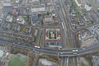 An aerial view of an urban area featuring numerous residential buildings, roads, parking spaces, and a railway line. The buildings exhibit modern architectural designs, predominantly with flat rooftops. A notable structure in the center appears to have a rooftop garden or recreational area. Roads are visible with a few vehicles, including buses and cars, moving through the area. Adjacent to the buildings, there are areas of green space, suggesting the presence of parks or lawns.