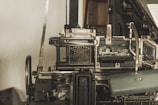 A vintage Linotype printing machine with metallic components and intricate mechanisms is displayed. The metal surface shows signs of wear, indicating its age and use. It features a plaque with text describing the model and manufacturer details.
