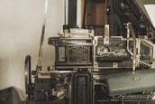 A vintage Linotype printing machine with metallic components and intricate mechanisms is displayed. The metal surface shows signs of wear, indicating its age and use. It features a plaque with text describing the model and manufacturer details.