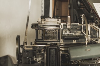 A vintage Linotype printing machine with metallic components and intricate mechanisms is displayed. The metal surface shows signs of wear, indicating its age and use. It features a plaque with text describing the model and manufacturer details.