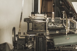 A vintage Linotype printing machine with metallic components and intricate mechanisms is displayed. The metal surface shows signs of wear, indicating its age and use. It features a plaque with text describing the model and manufacturer details.