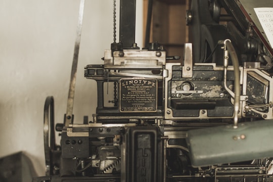 A vintage Linotype printing machine with metallic components and intricate mechanisms is displayed. The metal surface shows signs of wear, indicating its age and use. It features a plaque with text describing the model and manufacturer details.