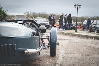 A vintage car with an open cockpit dominates the foreground, showcasing a detailed wooden steering wheel and metallic components. In the background, a group of people gathers near several parked modern cars. The setting appears to be an outdoor area with overcast skies and a streetlamp nearby.
