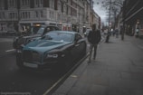 A luxury car, possibly a Rolls-Royce, is parked on a street lined with elegant, multi-story buildings. There is a black taxi cab nearby, and a person wearing a hooded jacket and beanie is walking on the sidewalk. The atmosphere is gray and somewhat moody, suggesting an overcast day in an urban setting.